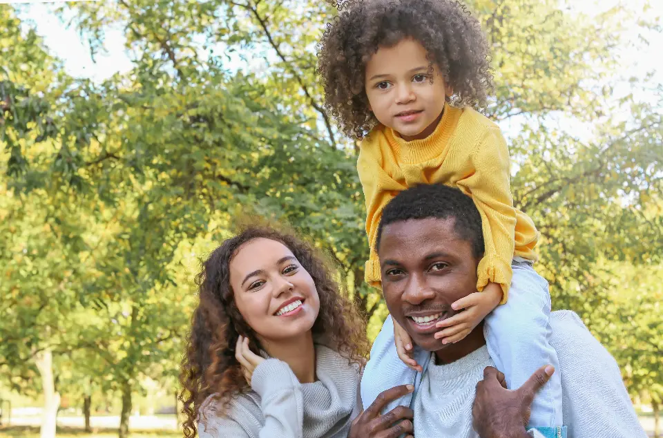 A parent carrying a young child on their shoulders beside another parent, all smiling, in a park