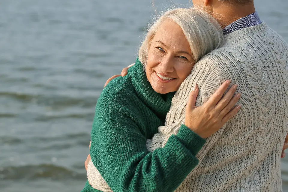 An older woman smiling over her partner’s shoulder as they embrace